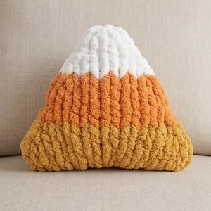 May include: A triangular-shaped plush pillow made of chunky yarn. The pillow features three horizontal color-blocked sections: a white top, an orange middle, and a yellow bottom, mimicking the appearance of a piece of candy corn.