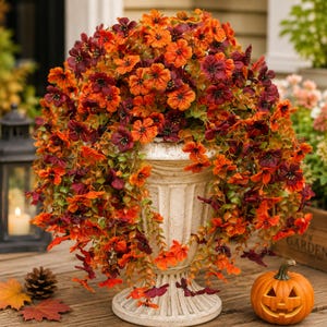 May include: A decorative urn filled with a dense arrangement of artificial flowers in shades of orange, deep red, and burgundy. The flowers feature small, five-petaled blooms and green foliage that cascades over the sides of the fluted white pedestal.
