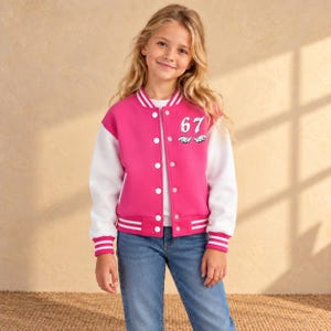 May include: A pink and white varsity jacket featuring long white sleeves, a snap-button front closure, and striped ribbed cuffs. The chest displays the number 67 in white with a small graphic detail underneath, paired with blue denim jeans.