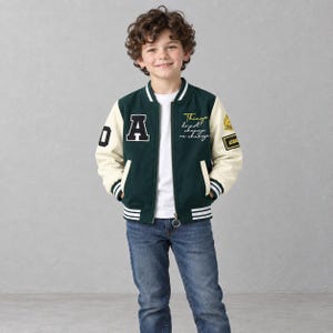 May include: A dark green and cream varsity jacket featuring a white letter A patch on the chest, striped ribbed cuffs, and yellow embroidered text that reads Things do not change we change, paired with a white t-shirt and blue denim jeans.