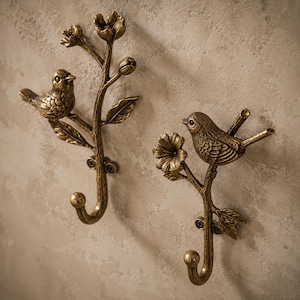 Peut inclure: Deux crochets muraux de couleur bronze. Chaque crochet représente un oiseau détaillé perché sur une branche avec des feuilles et des fleurs. Les crochets sont conçus pour suspendre des objets et ajouter une touche décorative.