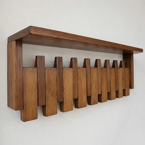 Puede incluir: Estante de madera montado en la pared con una serie de listones de madera verticales. El estante está hecho de madera de color marrón oscuro y tiene forma rectangular. Los listones están dispuestos en un patrón escalonado, creando un diseño único.