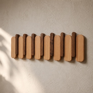 Op de afbeelding: Een houten wandkapstok met verticale houten latten met afgeronde toppen. De lamellen wisselen af tussen lichte en donkere houttinten, waardoor een decoratief patroon ontstaat. Ontworpen voor het ophangen van jassen, hoeden of andere items. (Ongeveer 60 cm breed).