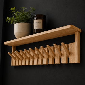Puede incluir: Un estante de pared de madera clara con una repisa superior horizontal y una fila de doce ganchos de madera inclinados debajo. Una pequeña planta verde en maceta y un frasco de vela de vidrio marrón con la etiqueta Sandalwood descansan arriba.