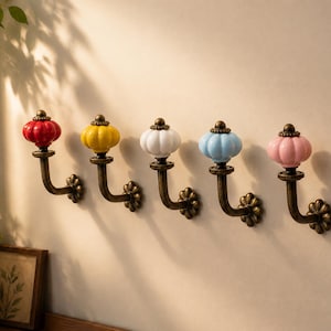 May include: Five decorative wall hooks with colourful ceramic knobs in red, yellow, white, light blue, and pink. The hooks have a bronze finish and a floral design at the base, mounted on a neutral wall.
