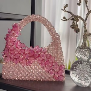 Può includere: Una borsa realizzata con perline rosa chiaro, dotata di un manico arrotondato e di una disposizione diagonale decorativa di perline traslucide a forma di fiore rosa scuro sulla parte anteriore, creando un aspetto floreale strutturato.