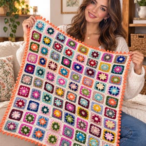 Op de afbeelding: Een vierkante gehaakte deken met een raster van veelkleurige granny squares met bloemmotieven in tinten roze, blauw, geel, groen en oranje. De deken is afgewerkt met een feloranje pomponrand en gebroken witte garenranden.