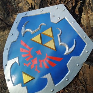 May include: A blue shield featuring a silver metallic border with visible rivets. The centre displays a red bird emblem positioned below a gold triangular crest. The shield has a pointed bottom and a notched top edge with a smooth, glossy finish.