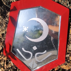 May include: A hexagonal mirror with a bright red frame. The reflective surface features a frosted white graphic design consisting of a crescent moon, a four-pointed star, and two symmetrical curved lines positioned below a small diamond shape.