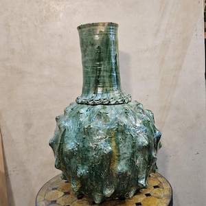 Può includere: Un grande vaso in ceramica sferico con un collo cilindrico alto. La superficie è ricoperta di punte sporgenti e rifinita con uno smalto verde lucido e screziato, caratterizzato da striature più scure e lievi sfumature marroni.