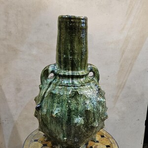 May include: A handcrafted, green ceramic vase with a textured surface and two handles. The vase has a bulbous body with a spiky texture and a tall, cylindrical neck. The color is a mottled green with darker accents, suggesting a rustic or antique aesthetic.