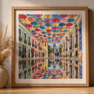 May include: A framed cross-stitch embroidery depicting a street scene with colourful umbrellas suspended overhead. The artwork features pastel-coloured buildings with balconies and flower boxes, reflected in a water-covered ground surface.