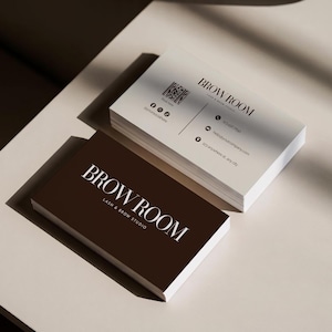Pode incluir: Uma pilha de cartões de visita retangulares para BROW ROOM LASH & BROW STUDIO. O cartão superior é castanho escuro com texto branco. A pilha inferior é branca com texto preto, incluindo um código QR, ícones sociais, telefone, email e morada.