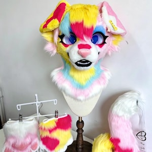Pode incluir: Uma máscara de cabeça de animal em pele sintética com um padrão multicolorido vibrante em amarelo, rosa e azul claro. A máscara tem olhos azuis grandes com pestanas pretas, um nariz rosa e boca aberta. Inclui patas e cauda em pele sintética.