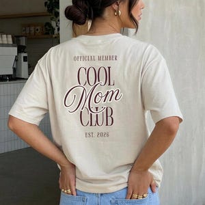 Könnte beinhalten: Ein cremefarbenes Kurzarm-T-Shirt mit dunkelbraunem Text auf der Rückseite, der OFFICIAL MEMBER COOL MOM CLUB EST. 2026 lautet. Das T-Shirt wird mit einer hellblauen Jeans kombiniert, die zwei Gesäßtaschen und eine sichtbare Taillennaht hat.