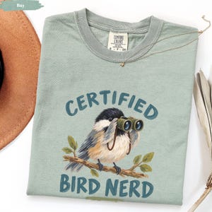 Peut inclure: Un t-shirt vert sauge avec une illustration d'une mésange à tête noire perchée sur une branche et regardant à travers des jumelles. Le texte au-dessus et en dessous de l'oiseau indique "CERTIFIED BIRD NERD" dans une police bleu foncé.