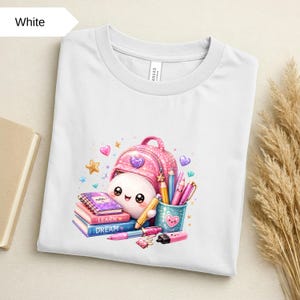Può includere: Una t-shirt bianca con la grafica di un simpatico personaggio bianco con uno zaino rosa glitterato, circondato da materiale scolastico tra cui un quaderno viola con la scritta notes, libri con learn e dream, matite e un bicchiere.