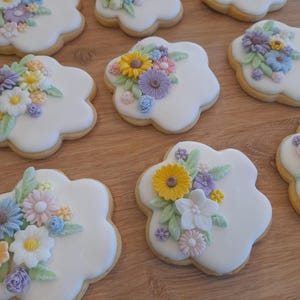 May include: Flower-shaped biscuits with white icing and colourful floral decorations. The biscuits feature yellow, purple, pink, and blue flowers with green leaves, arranged on a wooden surface.