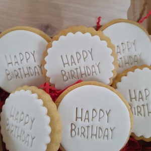 Op de afbeelding: Een verzameling ronde koekjes met een laagje gladde witte fondant, elk voorzien van de tekst HAPPY BIRTHDAY in reliëf. Sommige koekjes hebben een geschulpte rand terwijl andere glad zijn, allemaal verpakt in een doos met rode papieren snippers.