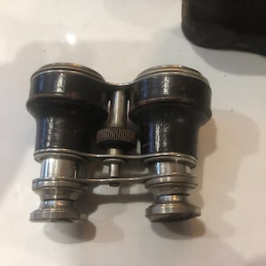 May include: Vintage opera glasses featuring black leather-covered barrels and silver-coloured metal detailing. The binoculars have a central focusing wheel and a classic design. A dark brown leather case is visible in the background.