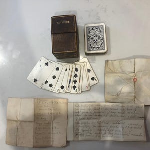 May include: An assortment of vintage playing cards, a brown leather card case with "PATIENCE" embossed on the top, and handwritten notes on aged paper. The cards are white with black suit symbols.