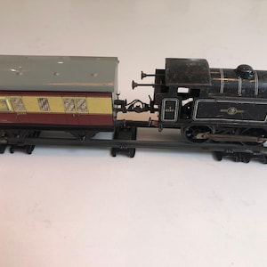 May include: A vintage toy train set consisting of a black steam locomotive with the number 82011 and a passenger carriage with a grey roof, yellow upper walls, and a maroon lower section, all positioned on a section of metal train tracks.