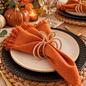 May include: A light-colored wooden napkin ring shaped like a pumpkin outline, holding a burnt orange linen napkin with frayed edges. The napkin rests on a speckled off-white plate, which is centered on a black ribbed plate and a woven placemat.