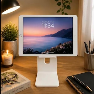 May include: A white tablet stand with a rectangular base and a support ledge, holding a tablet displaying a sunset landscape on its screen. The screen shows the text "Sexta-feira, 23 de maio" and "11:34" along with a "Pressione início para desbloquear" prompt.