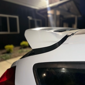 May include: A white car with a black spoiler. The spoiler is attached to the car's rear, with a black tape detail. The car's taillight is visible on the left side of the image. The background is a dark night scene.
