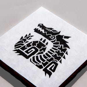 May include: A square tile featuring a black graphic print of a stylized dragon head and neck. The dragon design is composed of sharp, geometric shapes and angular lines, set against a white background on the top surface of the square object.