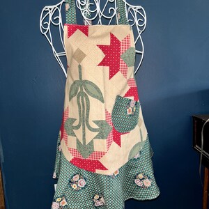 Peut inclure: Un tablier en tissu avec une partie supérieure beige ornée d'un motif de style patchwork à étoiles et fleurs rouges et vertes, incluant une petite poche. La partie inférieure est une jupe à volants à carreaux verts et blancs.