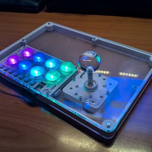 Puede incluir: Un mando arcade rectangular con panel superior de acrílico transparente, base blanca y joystick con cabeza esférica clara. El dispositivo incluye ocho botones circulares iluminados con luces LED de colores vibrantes morado, azul y verde.