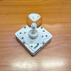 Pode incluir: Uma base de joystick arcade quadrada branca com um eixo metálico e um topo esférico que é transparente na metade superior e cor de âmbar na metade inferior. A base apresenta componentes eletrônicos expostos, parafusos e uma carcaça branca.