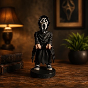 Può includere: Una statuetta di un personaggio che indossa una tunica nera con cappuccio e una maschera da fantasma bianca con la bocca spalancata. La figura ha mani visibili che tengono una piccola barra nera, indossa scarpe da ginnastica scure e poggia su una base nera.