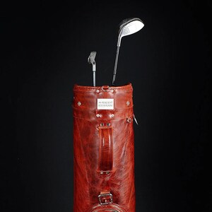 Może przedstawiać: Czerwono-brązowa sk&oacute;rzana torba golfowa z dwoma kijami golfowymi. Torba ma uchwyt i zapięcia na klamry. Na torbie widoczny jest napis "99 PERCENT HANDMADE".