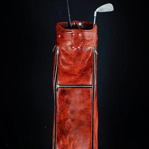 Może przedstawiać: Torba golfowa w stylu vintage, wykonana ze sk&oacute;ry w kolorze czerwono-brązowym. Posiada wiele przegr&oacute;dek zapinanych na zamek i srebrne okucia. Widoczne są dwa kije golfowe ze srebrnymi gł&oacute;wkami. Tło jest czarne.