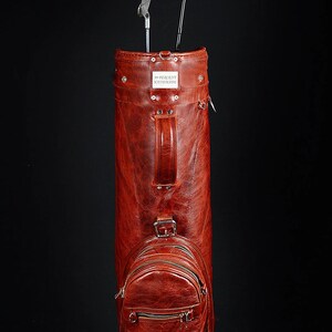 Może przedstawiać: Wysoka, czerwonobrązowa sk&oacute;rzana torba golfowa z dwoma kijami golfowymi. Torba ma uchwyt, małą kieszeń zapinaną na zamek i napis "99 PERCENT HANDMADE". Torba jest cylindryczna z teksturowaną powierzchnią.