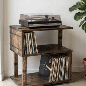 May include: A dark brown wooden storage unit with two open shelves for vinyl records, featuring metal accents and a black turntable with a clear plastic dust cover placed on the top surface. The unit holds multiple records.