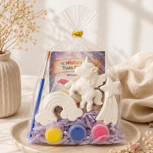 May include: A clear plastic gift bag containing white plaster figures of a unicorn, a rainbow, and a shooting star. The set includes a blue paintbrush, three small circular paint pots in yellow, blue, and pink, and a card labeled Mistura Tuas.