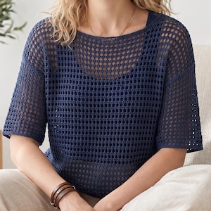 May include: A navy blue, open-weave knit top with short sleeves. The top has a square neckline and is worn over a darker blue underlayer. The person is wearing a brown leather bracelet. The person is sitting down.