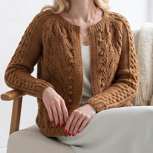 May include: A long-sleeved, open-front cardigan in a warm brown hue, featuring intricate cable knit patterns, textured bobble details, and ribbed cuffs. The garment has a round neckline and a structured knit design throughout the sleeves and body.