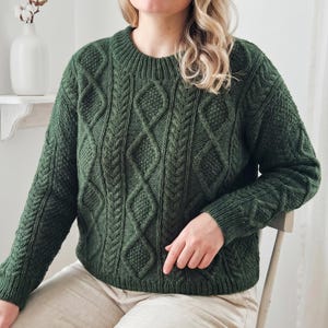 May include: A dark green knit sweater with a crew neck and intricate cable knit patterns. The sweater features diamond and braided textures, with ribbed cuffs and hem. The sleeves are long, and the overall style is classic and cozy.