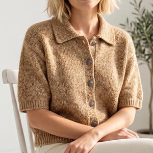 Peut inclure: Cardigan à manches courtes de couleur beige moucheté, avec un col et des boutons décoratifs. La personne porte le cardigan en étant assise sur une chaise blanche. Vêtement de style décontracté.