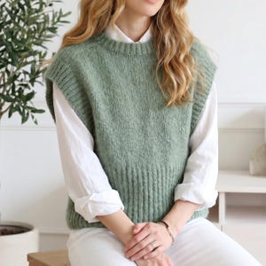 May include: A sage green knitted vest with a crew neck. The sleeveless jumper is worn over a white collared shirt. The model is wearing white trousers and has blonde hair. The vest is a soft, textured knit.