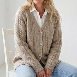 May include: A beige cable-knit cardigan with a V-neck and three button closures. The sweater features a textured pattern on the sleeves and front panels. The person is wearing a white collared shirt and blue jeans.