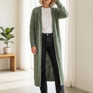 May include: A long, sage green cardigan with two front pockets. The cardigan is worn over a white t-shirt and black jeans. The outfit is styled in a well-lit room with a plant and wooden table in the background.