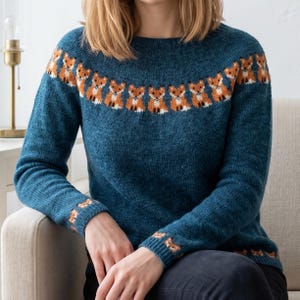 Puede incluir: Jersey azul turquesa con un patrón repetido de zorros naranjas y blancos alrededor del cuello y los puños. El punto de manga larga tiene cuello redondo y está hecho de un material suave y texturizado. El jersey lo lleva una persona sentada en un sofá beige.