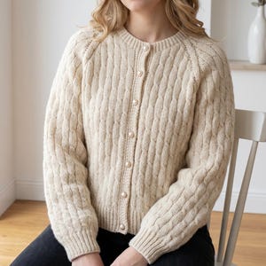 May include: A cream-coloured cable-knit cardigan with a round neckline and long sleeves. The button-down sweater features pearl-like buttons and a textured pattern. The garment is made of a soft, warm material.