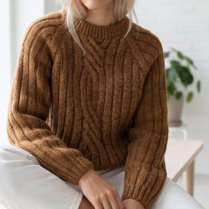 May include: A long-sleeved, crew-neck sweater in a rich, warm brown color. The garment features a prominent vertical cable knit pattern running down the center, flanked by vertical ribbed textures on the sleeves and sides of the torso.