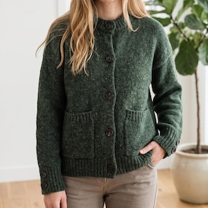 Op de afbeelding: Een donkergroen vest met een ronde hals en knoopsluiting. Het heeft twee voorzakken en donkerbruine knopen. De mouwen hebben geribde manchetten met knopen. Het model draagt een beige broek.